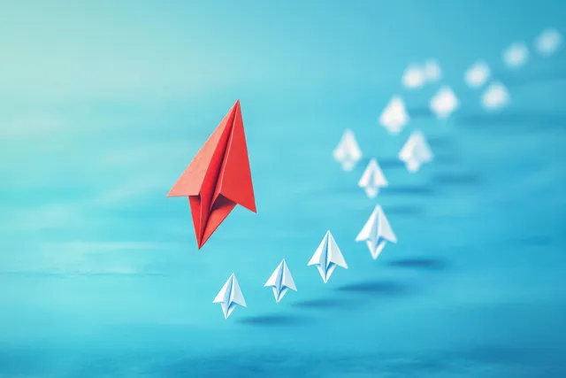 Red paper airplane leads a line of white planes with trailing motion on blue backdrop