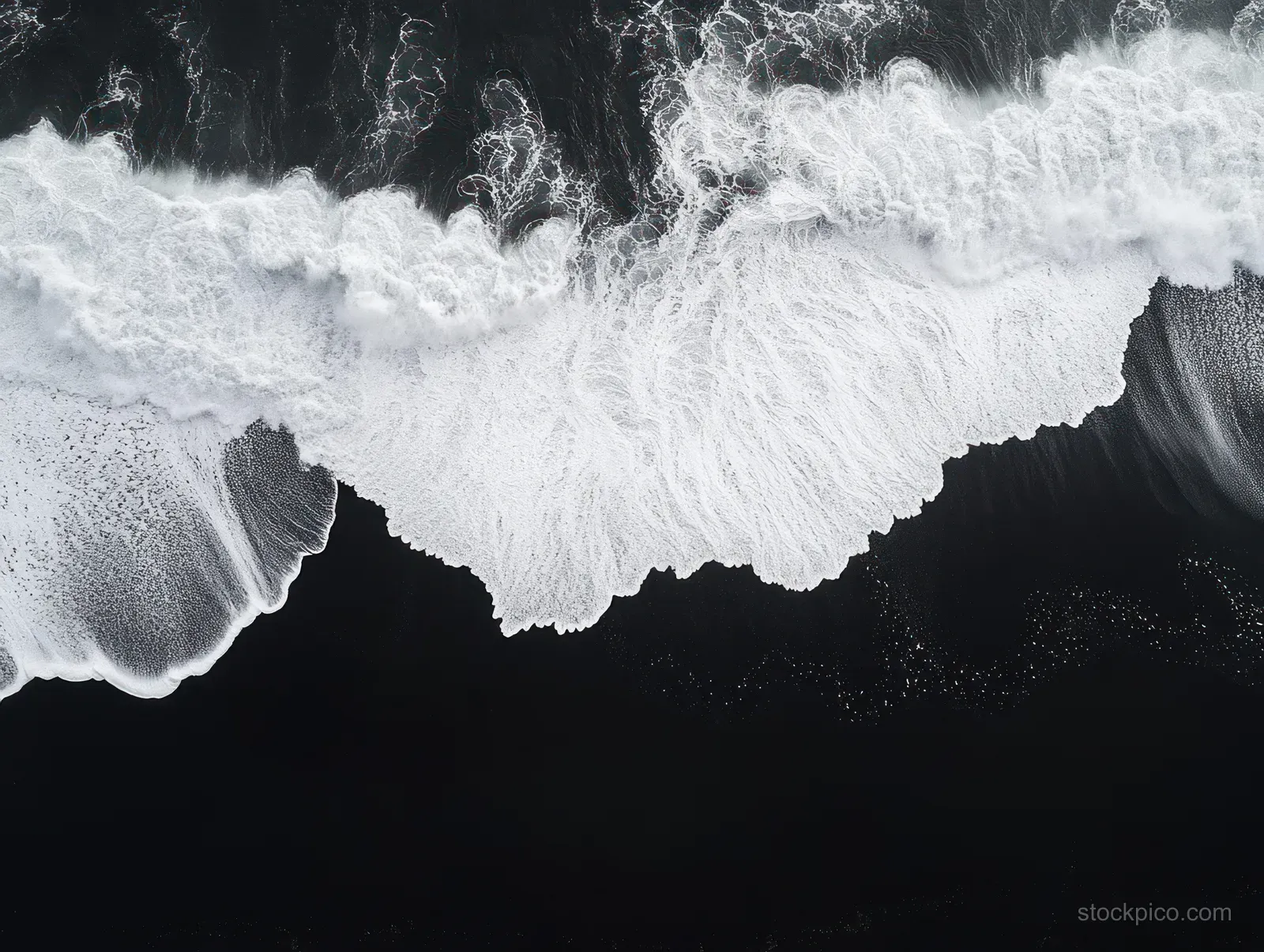 Aerial photo shows white foam waves breaking on dark sandy beach with sharp textured wave patterns.