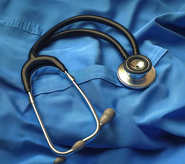 Black-and-silver stethoscope lying on blue medical scrubs fabric with the chest piece and tubing visible.