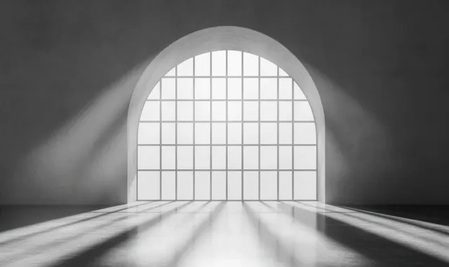 Sunlight streams through an arched grid window, casting long shadows on an empty smooth floor inside a minimalist room.