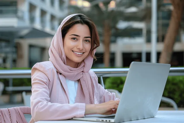 Smiling woman in a pink jacket uses a laptop outdoors at a modern city plaza surrounded by greenery.