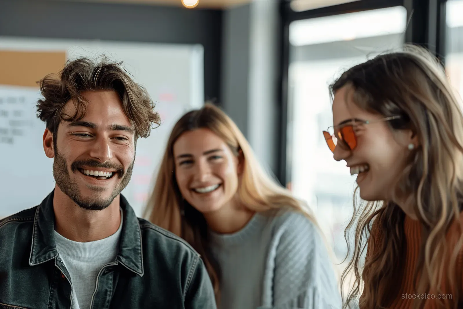 Three coworkers smile during a meeting in a modern office, with coworkers and a whiteboard blurred behind them.