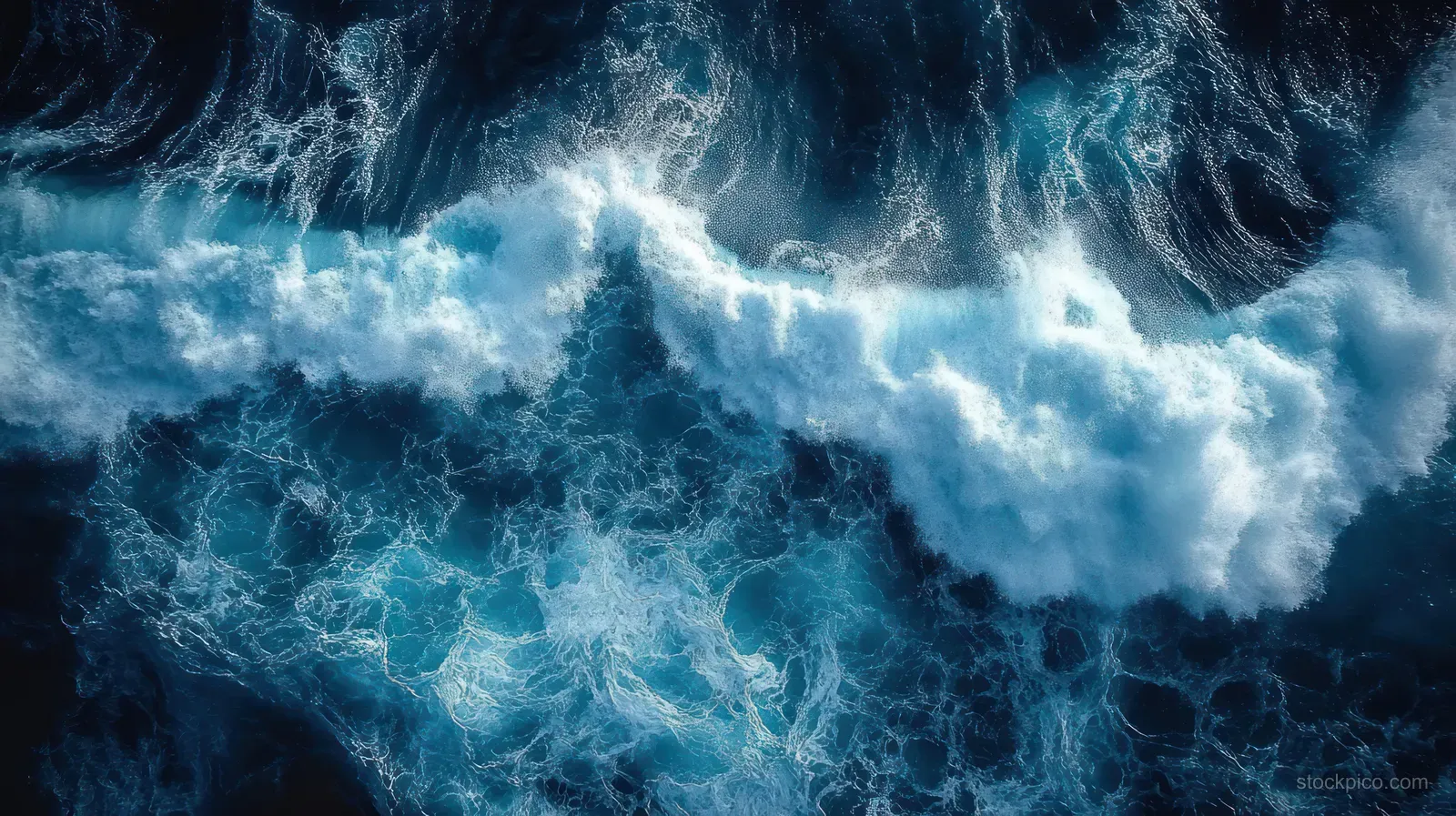 Aerial view shows ocean waves crashing and foaming over deep blue water with visible spray and turbulence.