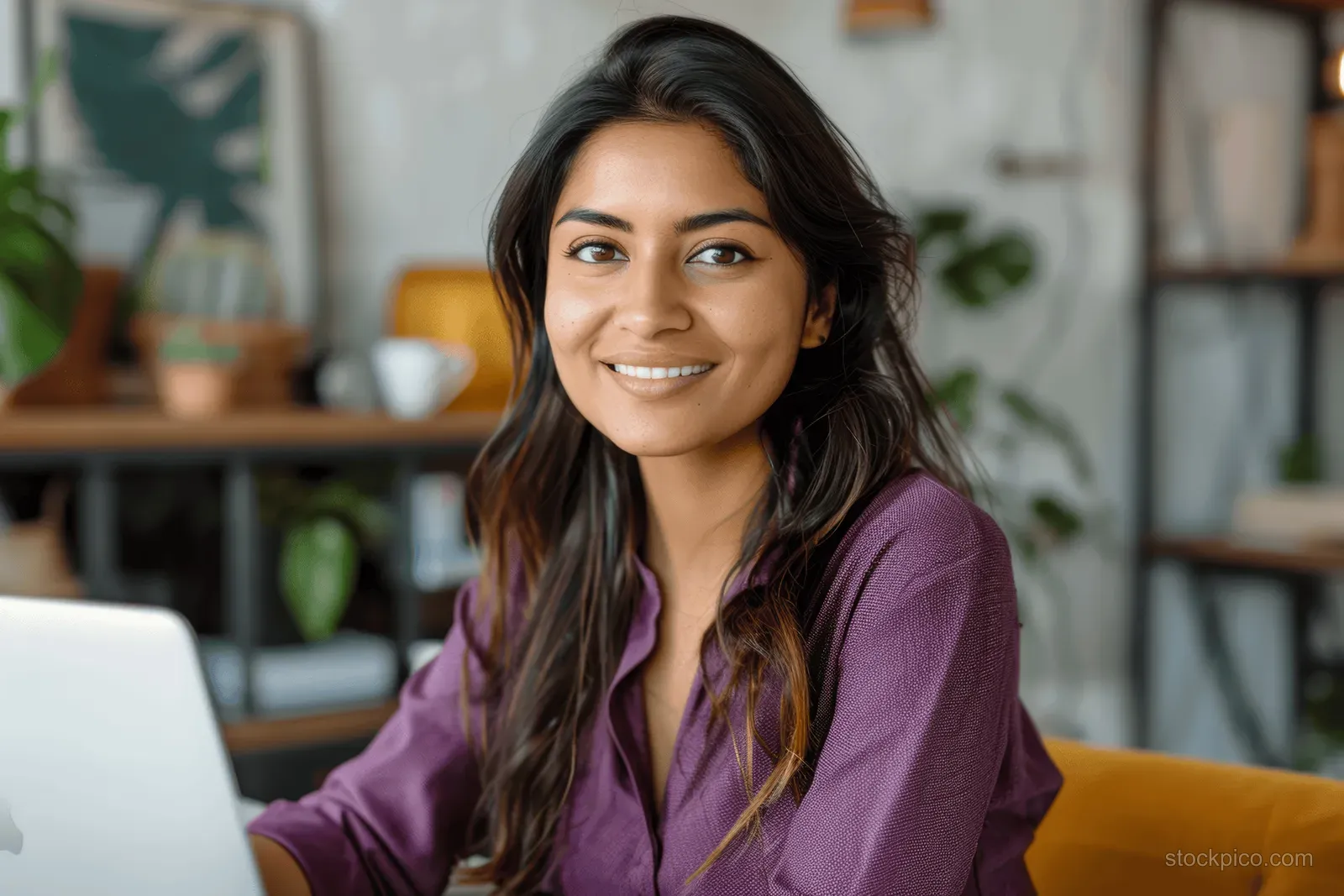 Cheerful Indian woman in a purple shirt smiles at the camera while working on a laptop at her desk.