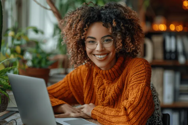 A smiling woman in an orange sweater takes notes on a laptop while watching a webinar at a home office desk.