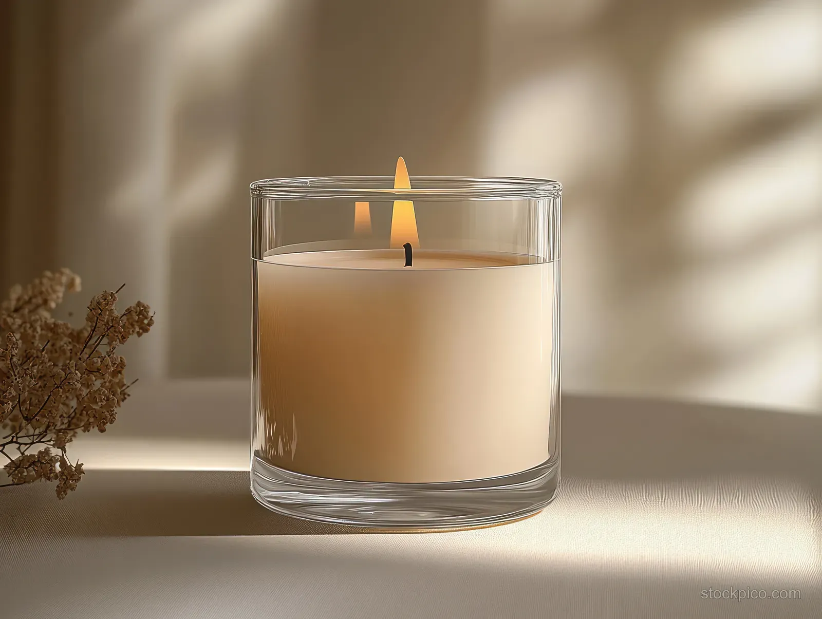 A lit candle in a clear glass holder with dried flowers beside it, casting soft shadows in warm natural light.