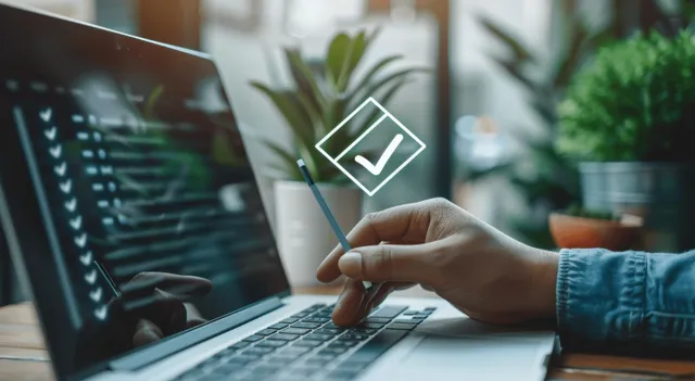 A hand holds a stylus near a laptop screen with a check mark icon.