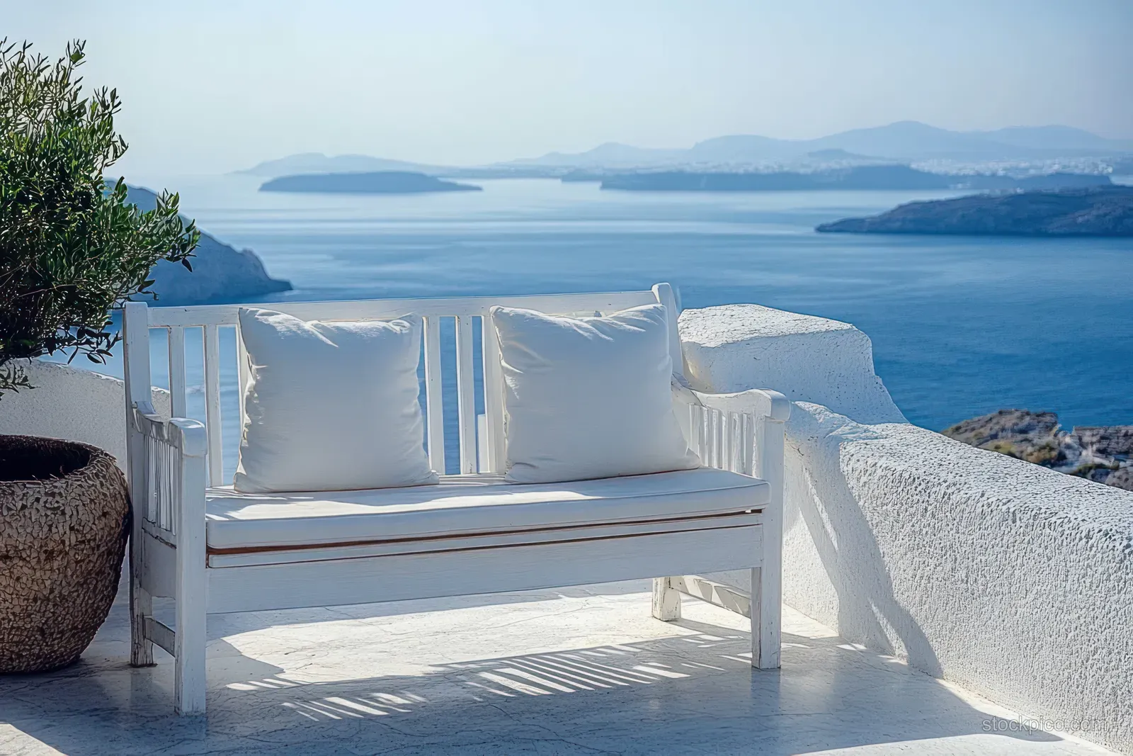 White bench with cushions on a terrace overlooks calm blue sea and distant islands under clear sky.