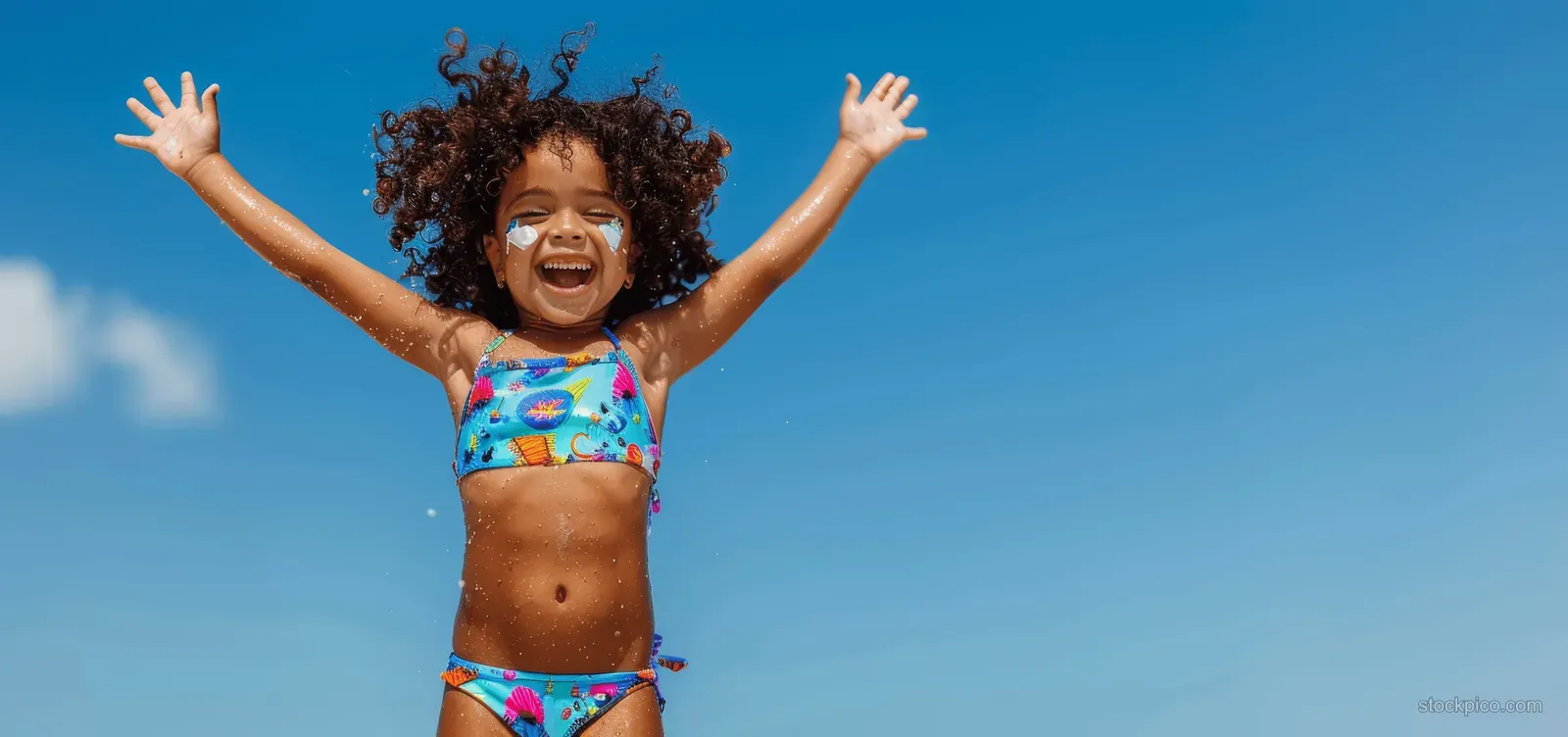 A joyful girl jumps on a sunny beach with arms raised, wearing a colorful swimsuit beneath a clear blue sky.