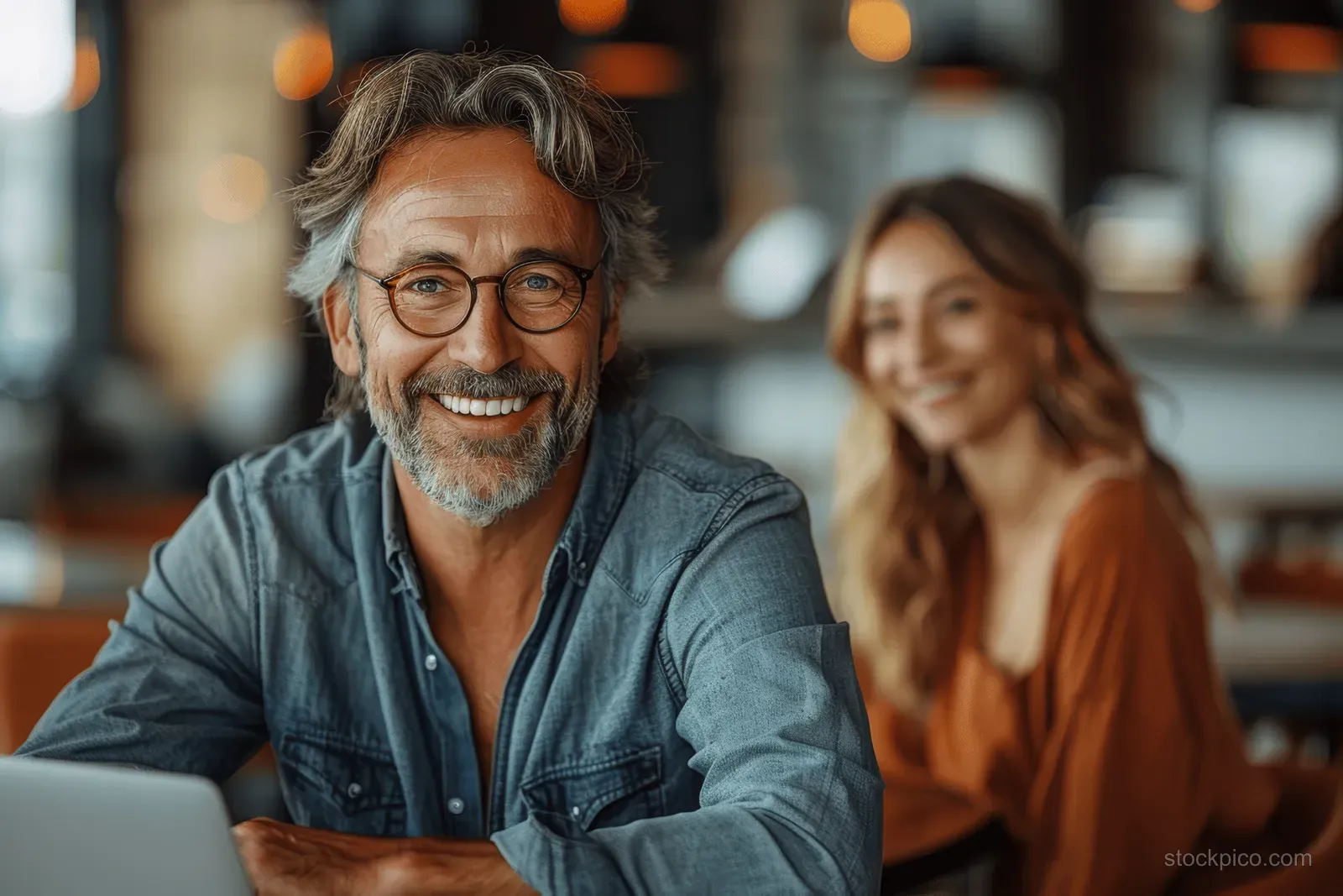 Glasses-wearing businessman types on a laptop while a female colleague listens during an office meeting.