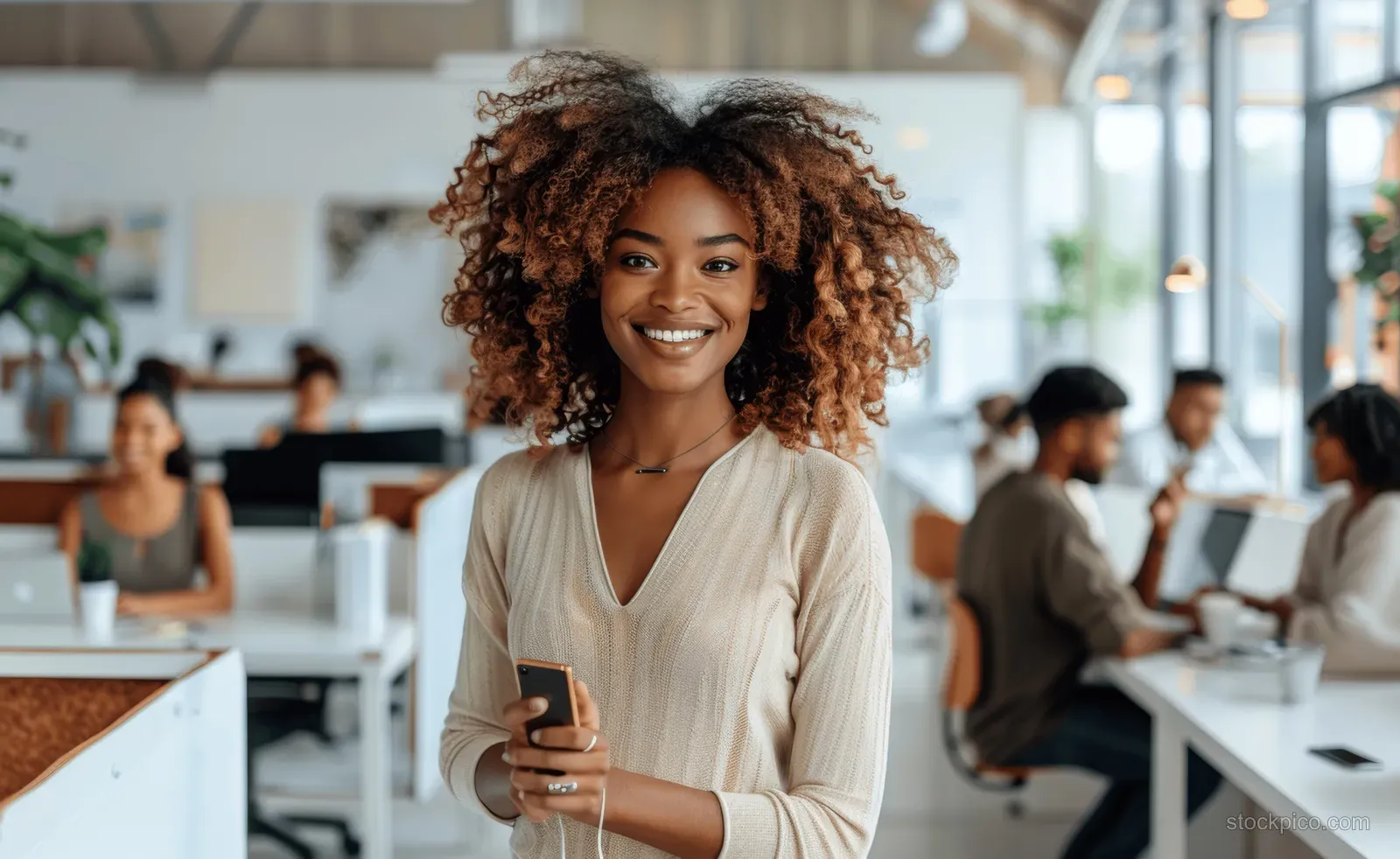 Smiling African American woman holds a smartphone in a bright office with coworkers behind her.