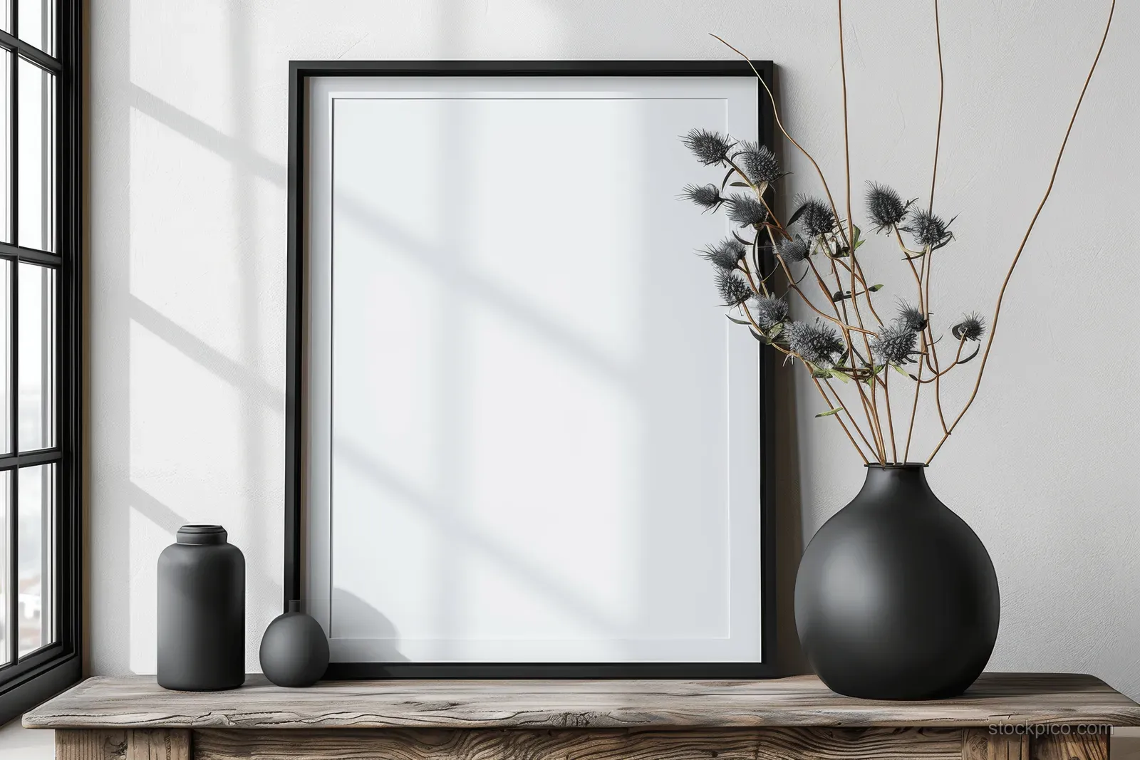 Minimalist black frame mockup on a wooden console with a blank white poster and a thistle-filled vase.