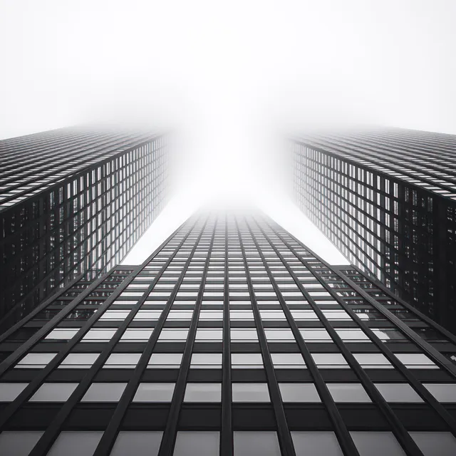 Three tall skyscrapers frame a foggy skyline from a ground-level upward angle with strong vertical lines.