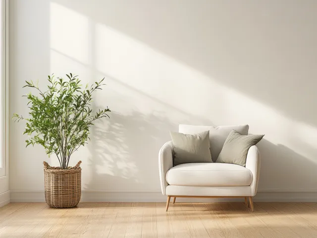 Sunlit minimalist living room corner with a white armchair, soft pillows, and a green plant in a wicker basket on wooden floor.
