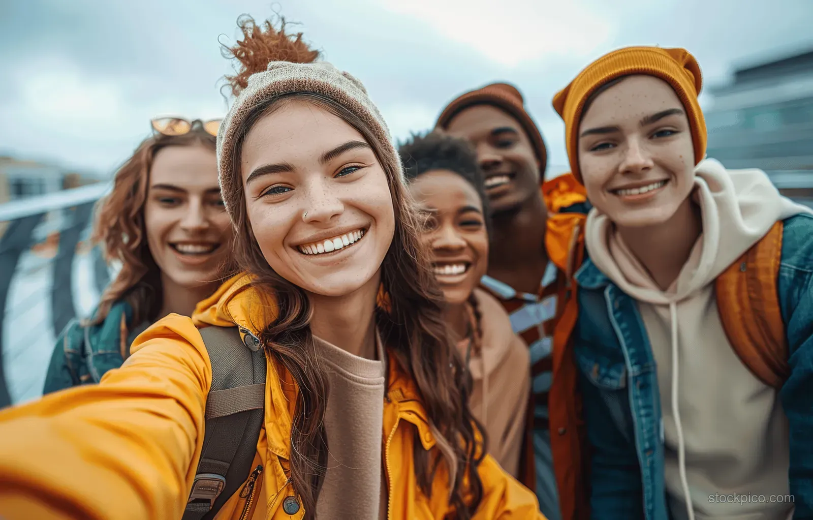 A diverse group of friends smiles for a selfie on a modern city bridge while standing outdoors.
