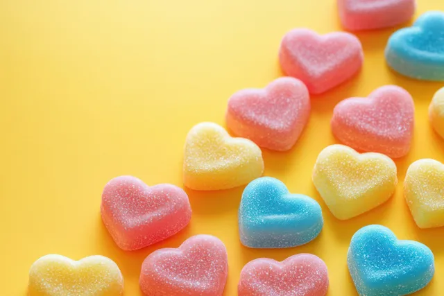 Colorful heart-shaped gummy candies with sugar coating scattered on a bright yellow background.