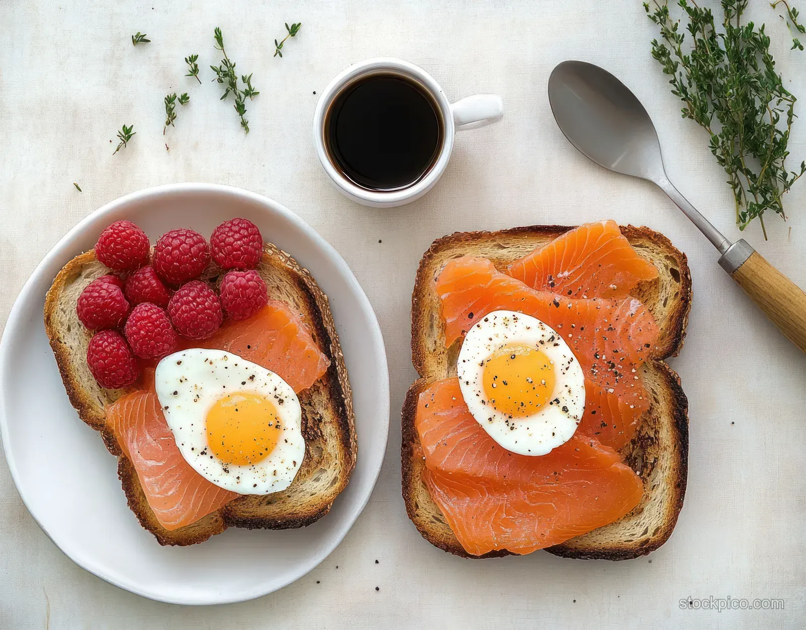 Two toast slices topped with smoked salmon and sunny-side-up eggs on white plates, with raspberries and black coffee beside.