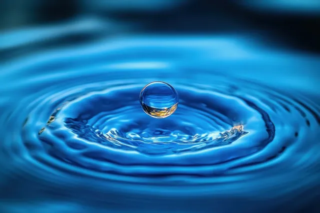 A clear water droplet falls into a blue water surface, creating concentric ripple rings.