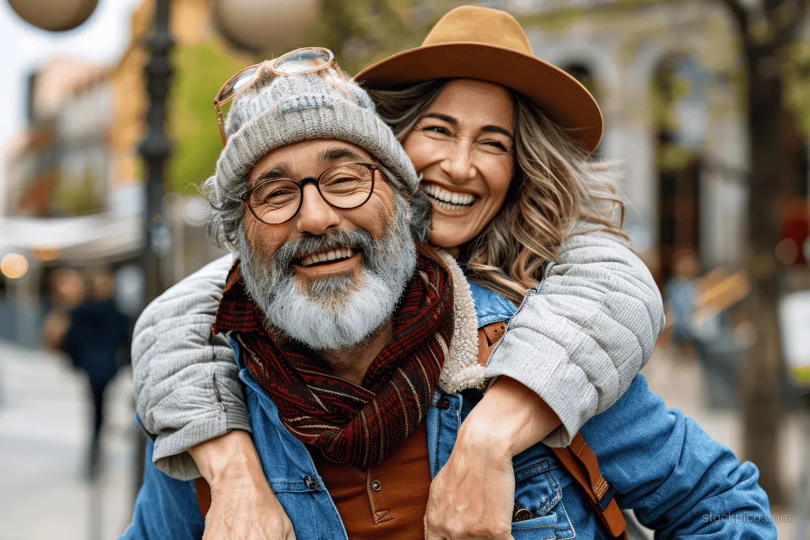 A senior man piggybacks his partner through a city square while they smile and wear winter hats and scarves.