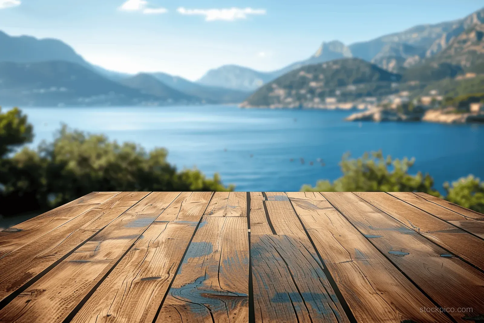 Rustic wooden table overlooking a calm lake with mountains and green trees under a blue sky.