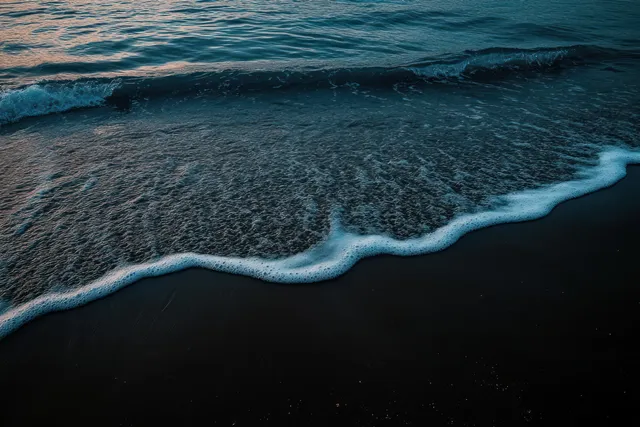 Ocean waves roll onto a dark sandy beach with delicate foam lines under a dim twilight sky.