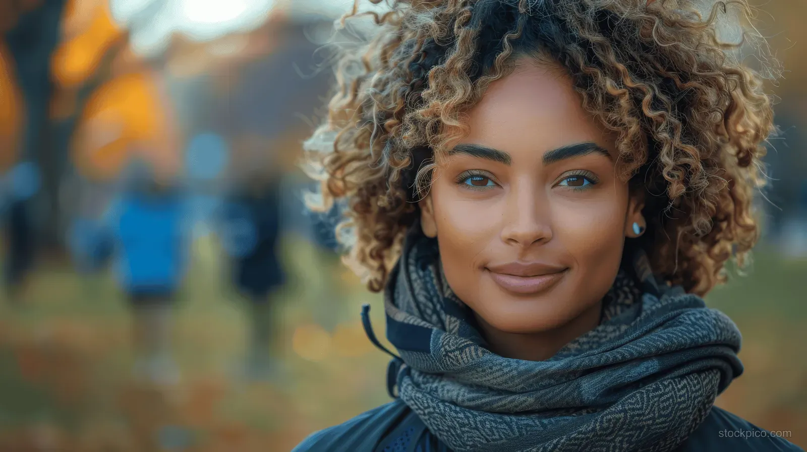 A smiling woman with curly hair stands outdoors with a scarf in a blurred park.