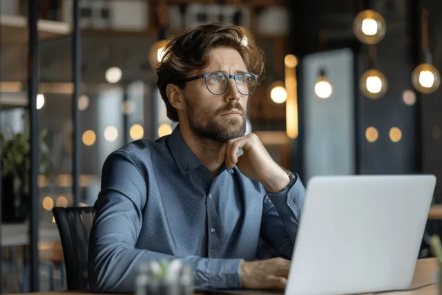 Thoughtful Businessman with Glasses Working on a Laptop in a Modern Office