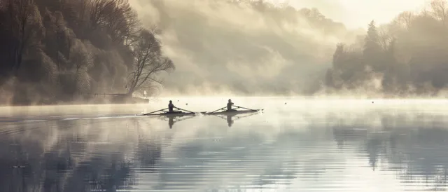 Women row a dual sculls boat across a misty lake at dawn as the water mirrors their silhouettes.