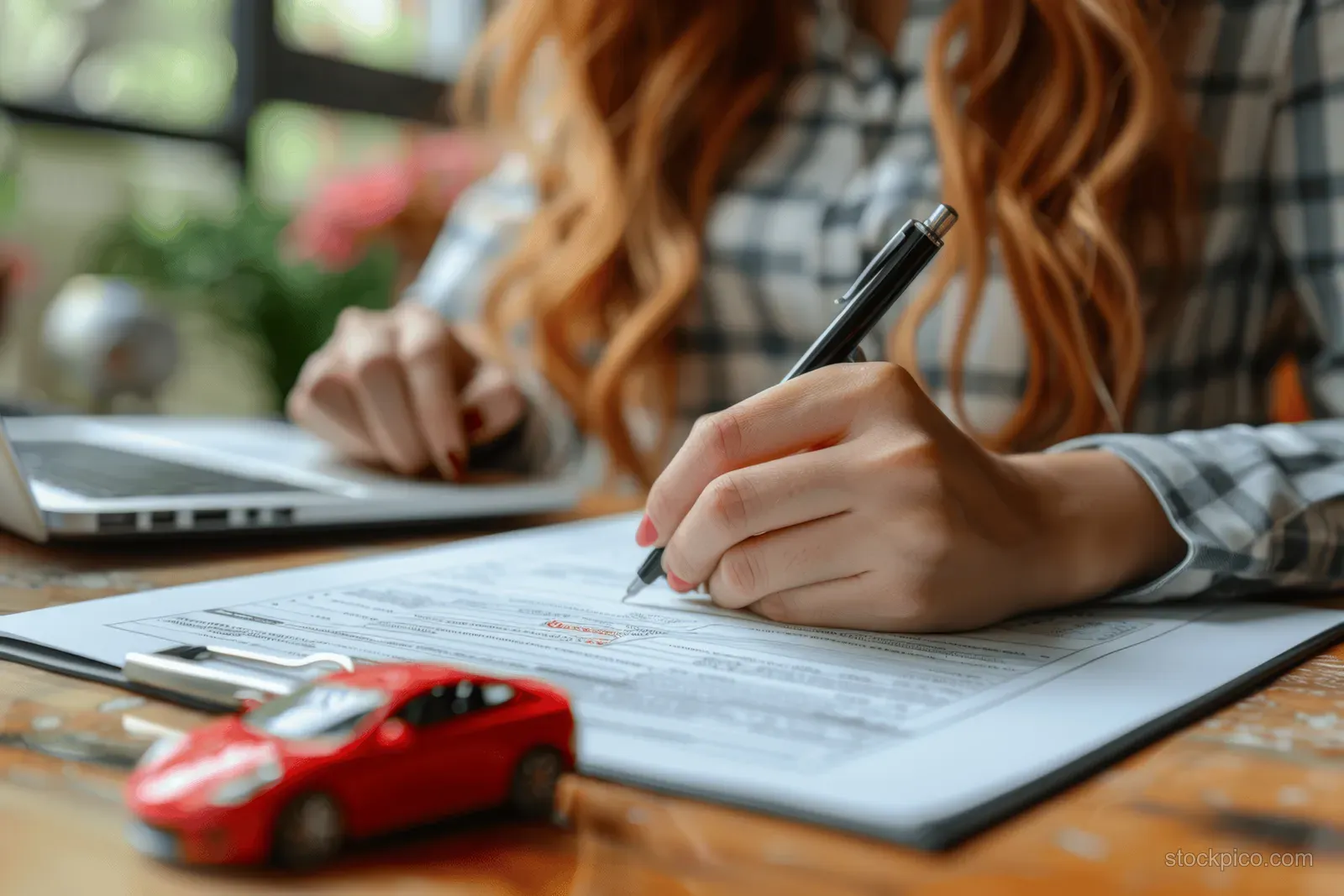 Woman signs car paperwork at a desk while a toy red car sits beside an open laptop.