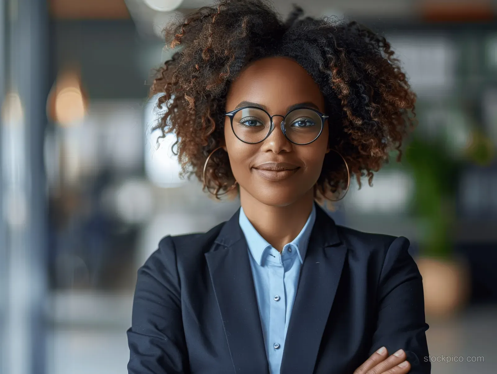 Smiling Black businesswoman with curly hair and glasses stands confidently in a modern office wearing a professional suit.