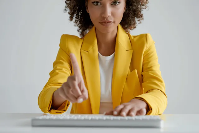 A woman in a yellow blazer types on a laptop keyboard at a white desk in a minimal office.