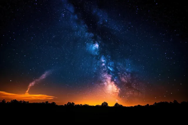 Milky Way brightens a starry night sky above dark tree silhouettes and orange horizon glow
