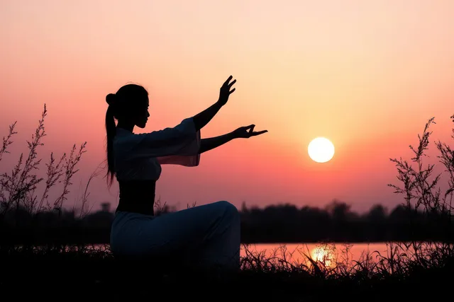 Silhouette of a woman practicing tai chi beside a calm lake at sunset under orange and pink sky.