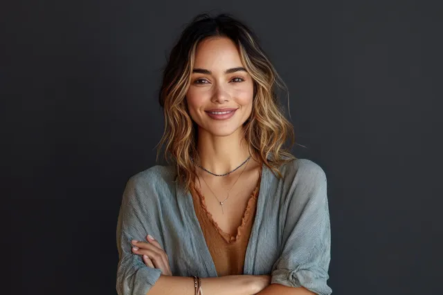 A woman smiles with her arms crossed in a centered studio portrait against a dark gray background.