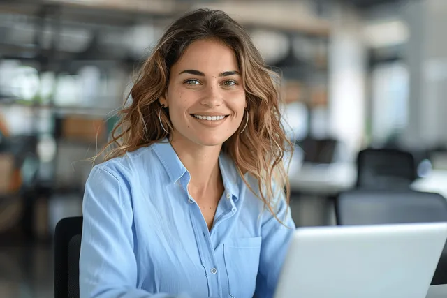 Smiling businesswoman in blue shirt working on laptop in modern office, looking away