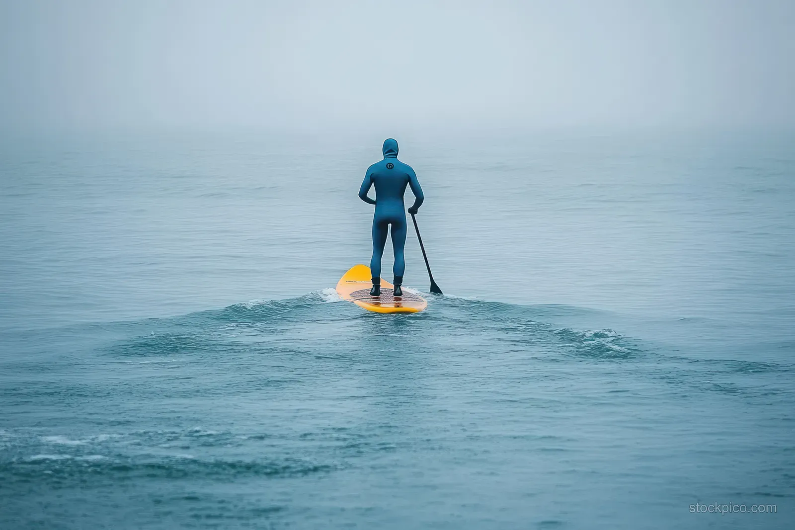 A lone paddler in a wetsuit stands on a stand-up paddleboard on misty calm ocean water.