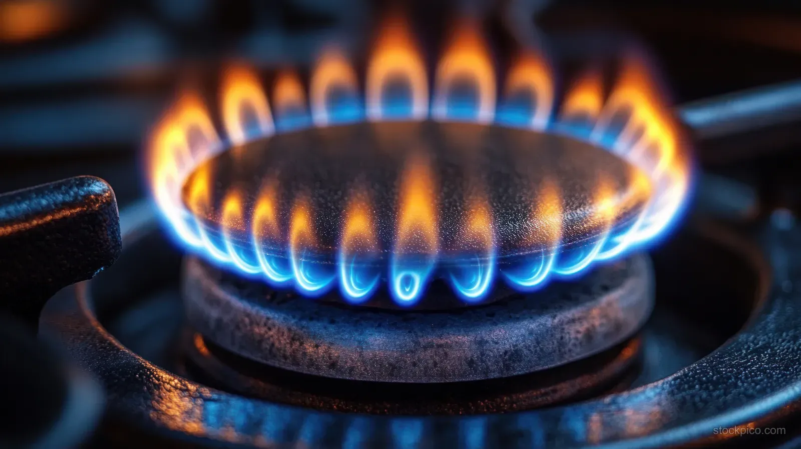 Blue and orange gas flame burns steadily around a stovetop burner ring on a metal stove in dim kitchen light.