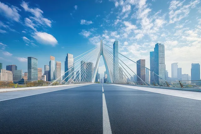 An empty suspension bridge with white cables stretches toward a modern city skyline under a bright blue sky.