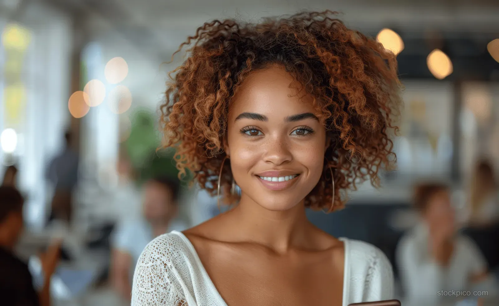 Smiling young black businesswoman holding a smartphone in a modern office with coworkers in the background.