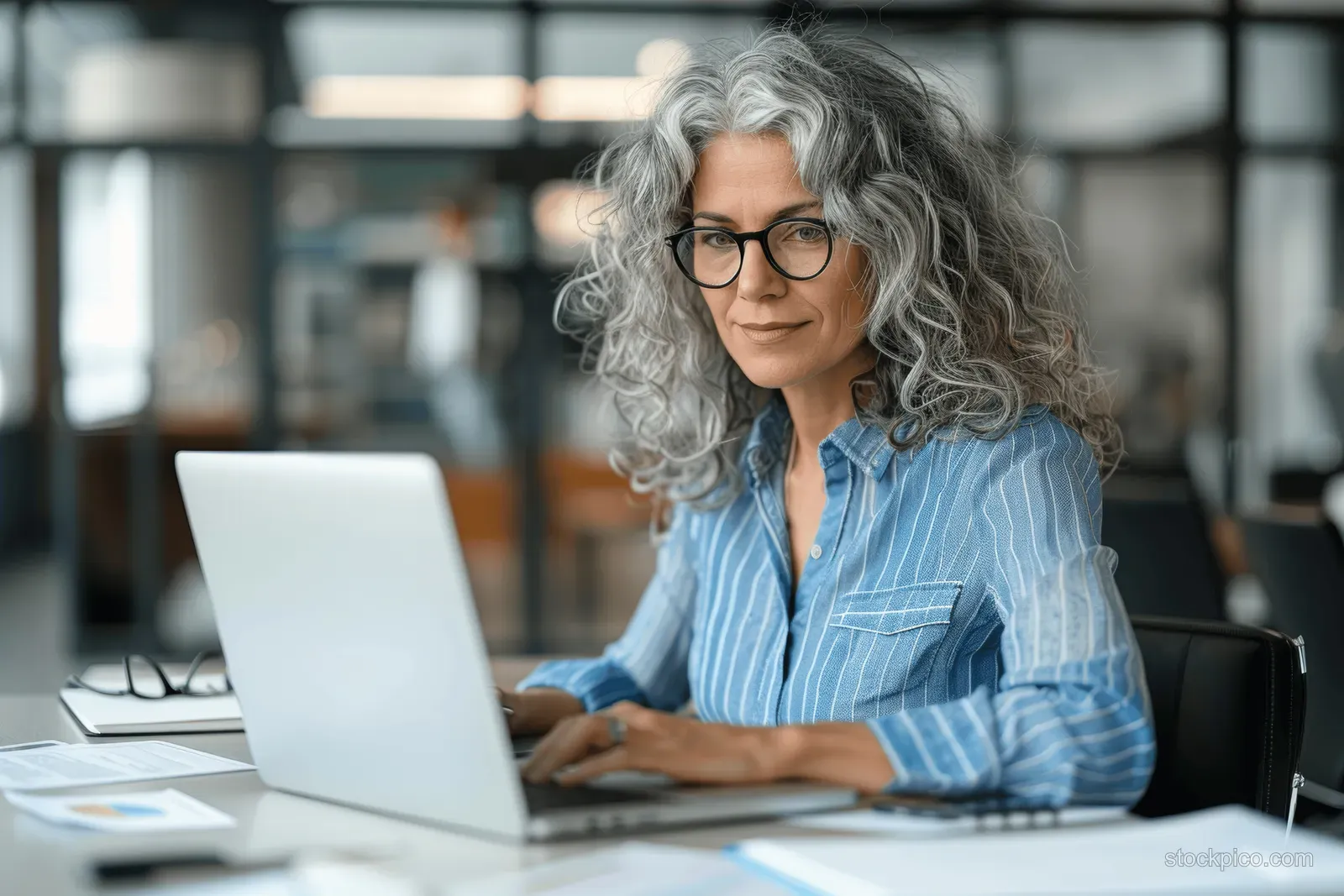 Curly gray-haired manager in glasses types on a laptop at a modern office desk with documents.