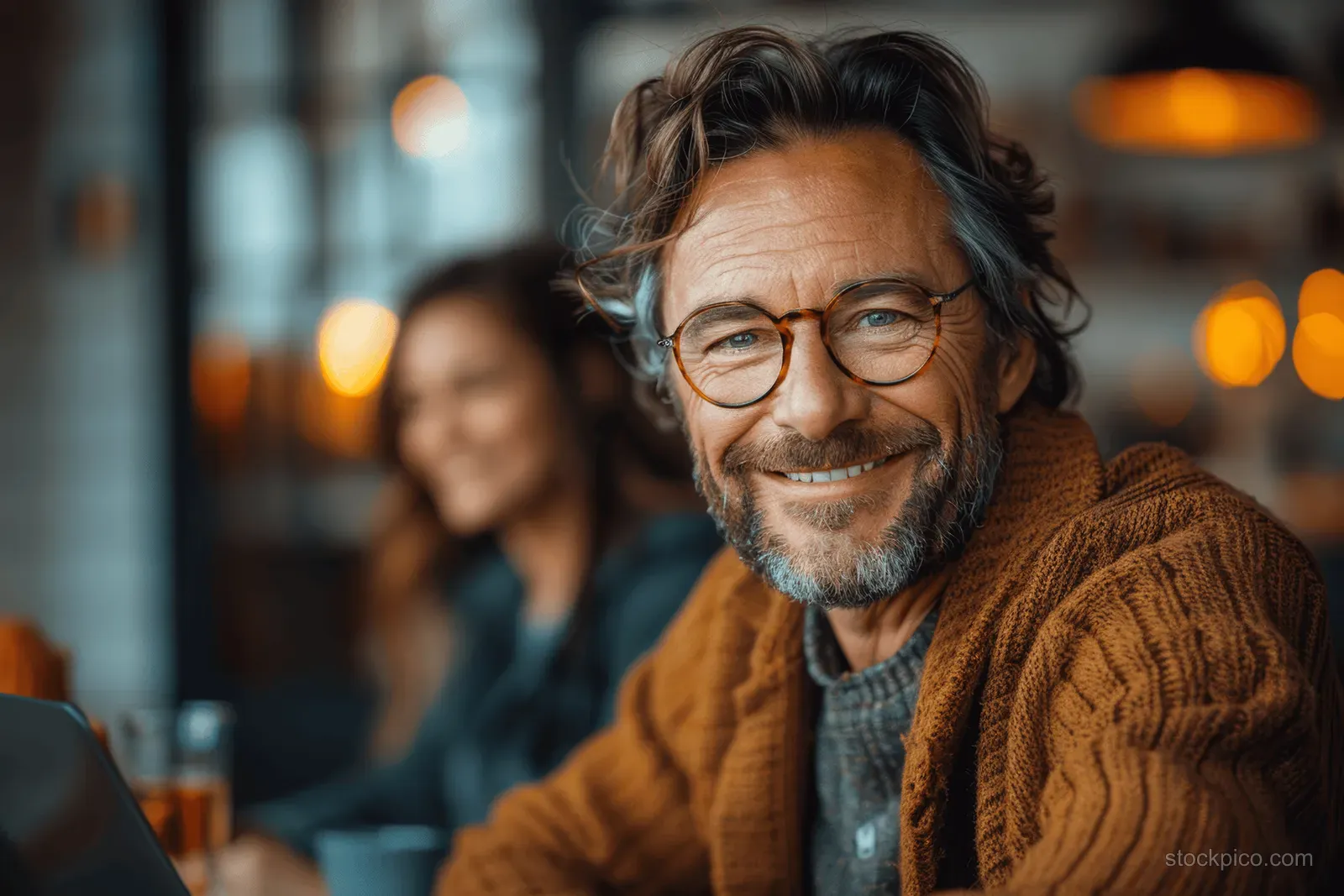 Cheerful man with round glasses sits at an office desk working on a laptop during an online meeting.
