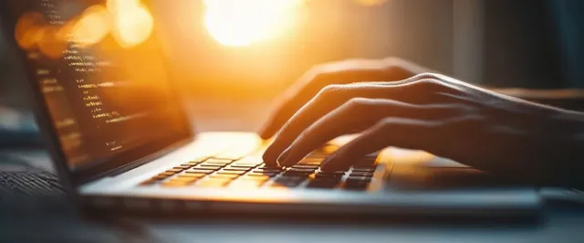 Hands type on a laptop keyboard in warm golden sunlight with soft lens flare across the desk.