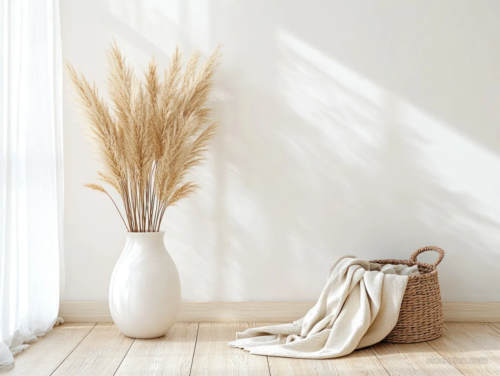 Pampas grass stands in a white vase beside a woven basket and folded blanket on a wooden floor.