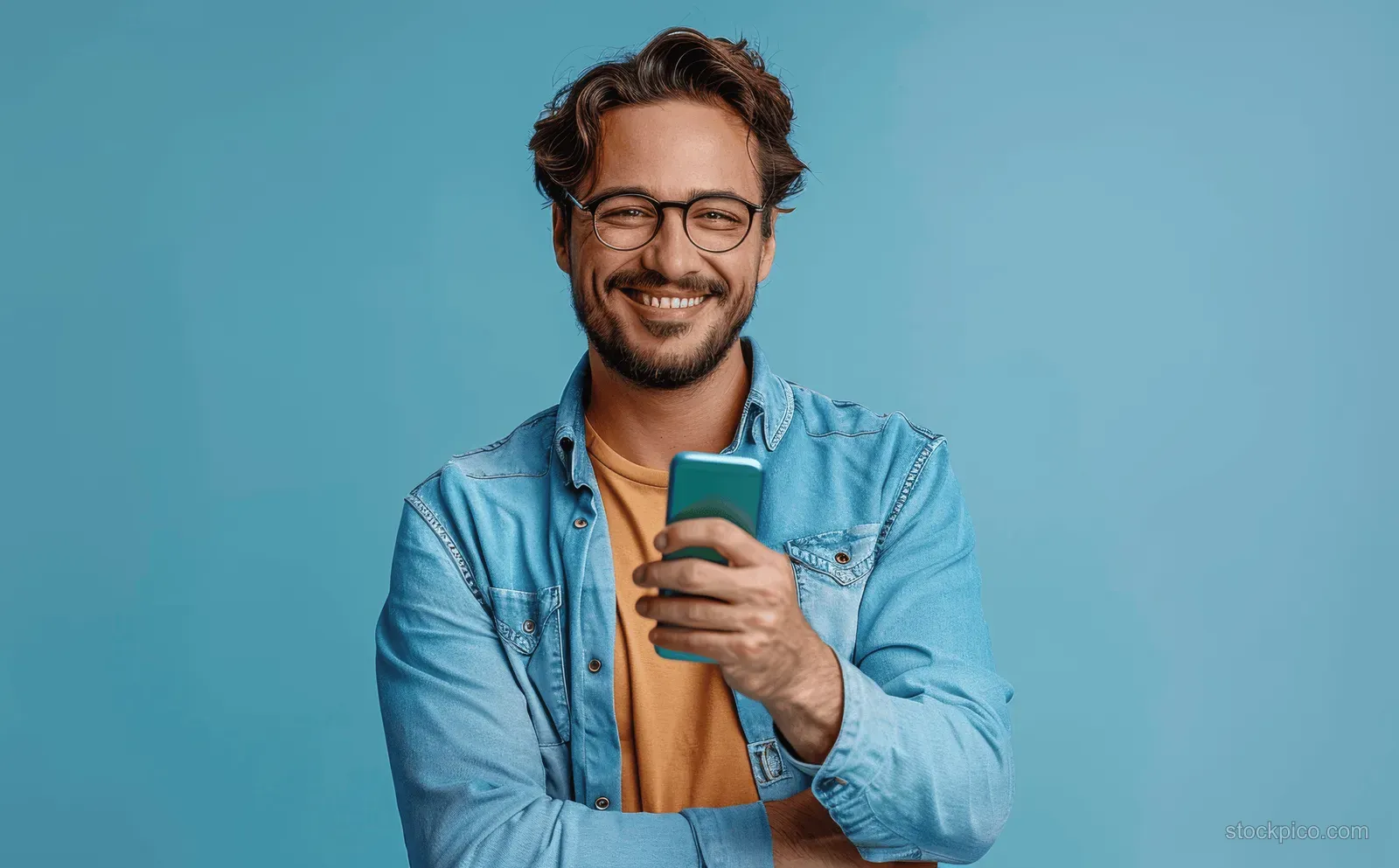 Smiling man in casual jacket holds a smartphone while standing against a solid blue background.