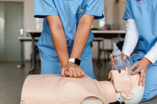 Two healthcare professionals practice CPR and ventilation on a resuscitation mannequin with a bag-mask in a clinical training room.