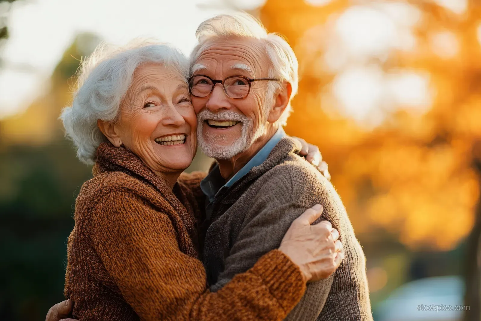 An elderly couple hugs and smiles outdoors at golden hour with warm autumn trees behind them.