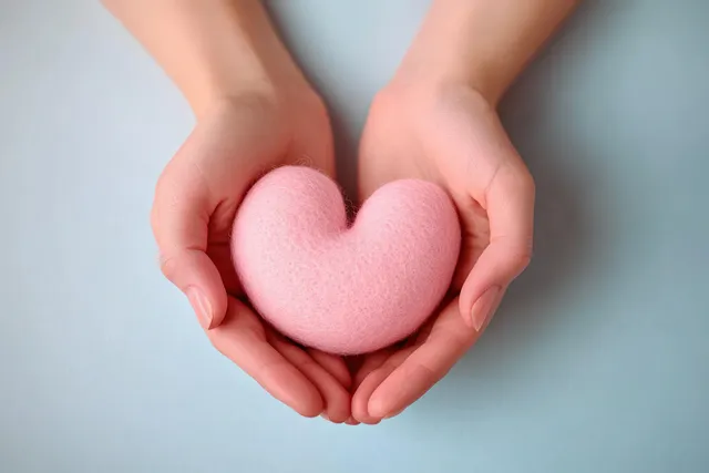 Two hands cradle a soft pink heart on a light blue background, showing care