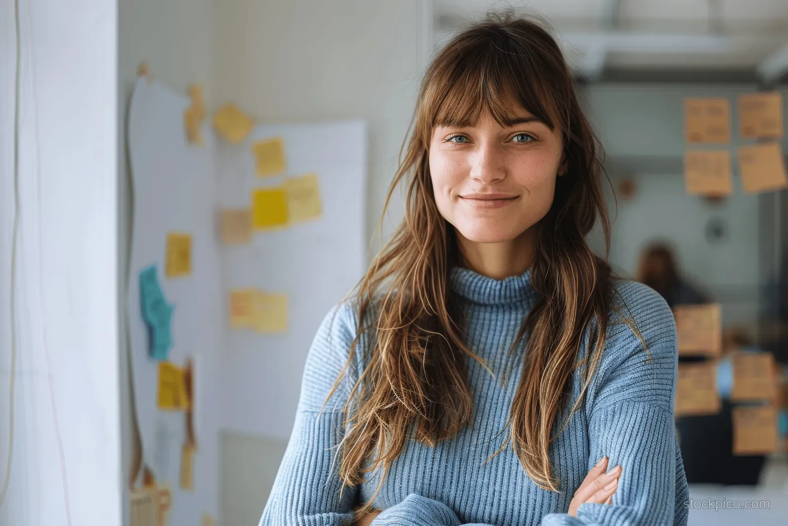 A woman in a blue sweater stands with arms crossed in a creative office with sticky notes on walls.