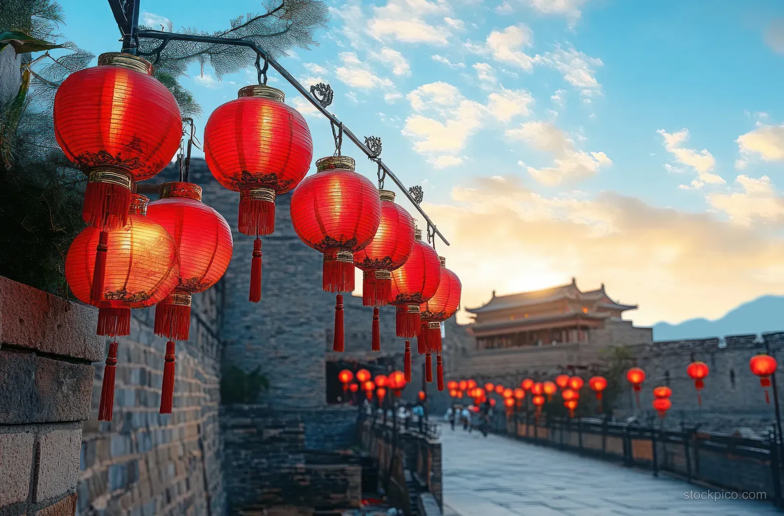 Red traditional lanterns hang along an ancient stone wall walkway beside a fortress at sunset with clouds overhead.