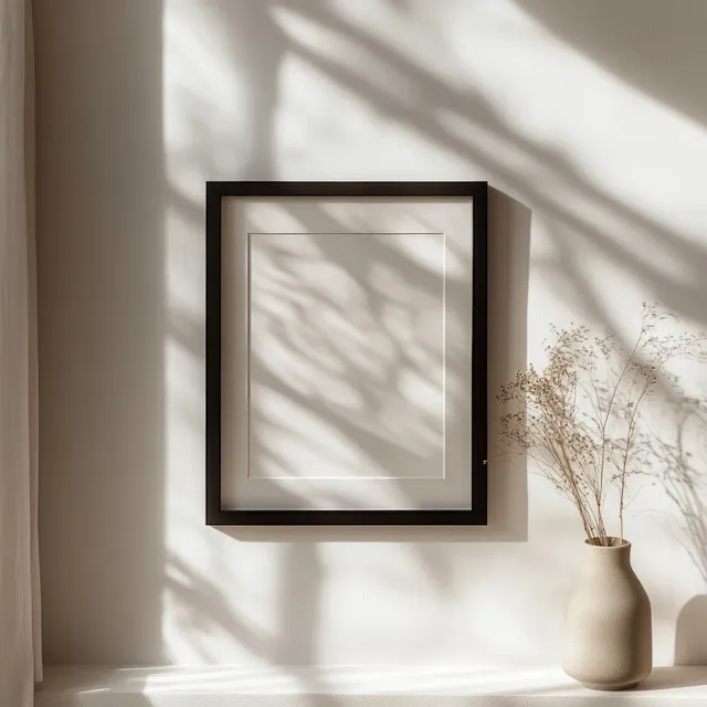 A blank black picture frame casts soft shadows on a wall beside a beige vase with dried branches.